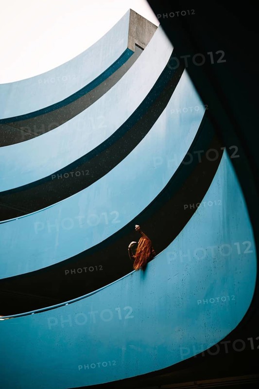 An unrecognizable woman with vibrant red hair stands at a balcony of a blue parking lot. The unique architectural details of the building create a contrast with her hair color, making for an eye-catching scene