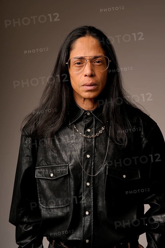 Close-up portrait of a Latin man wearing a black leather shirt and glasses, posing confidently. The neutral background enhances the subject's engaging expression, highlighting his style