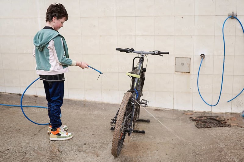 A young boy cleans a muddy mountain bike with a blue hose against a tiled wall. The scene captures responsibility and outdoor play on a concrete surface