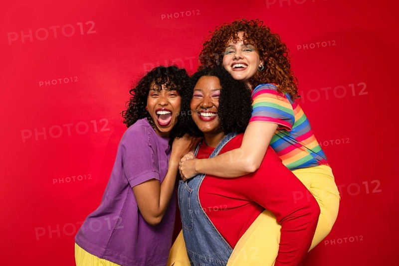 Three friends joyfully embrace, exuding happiness and warmth against a vibrant red background. Their bright and colorful clothing enhances the cheerful atmosphere
