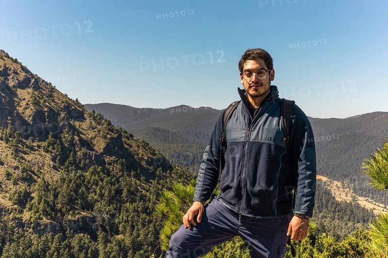 A man with a backpack stands confidently on a mountain trail, surrounded by lush, forested hills under a bright, clear sky A perfect image of outdoor adventure and exploration