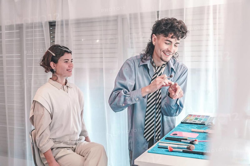 Young make-up artist preparing brushes and testing colors while applying makeup on a female model sitting beside him in a bright, well-lit studio filled with creativity