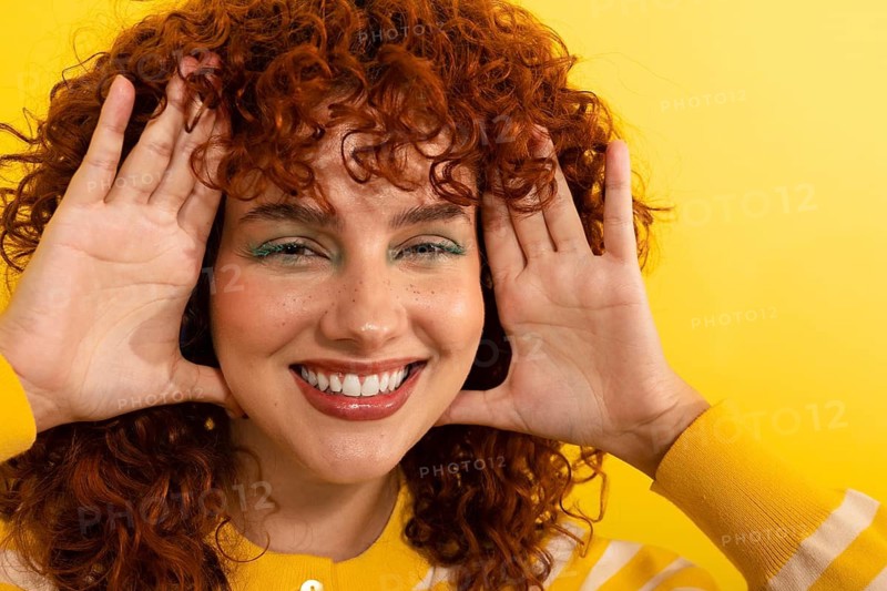 A smiling woman with curly red hair poses playfully against a bright yellow background, highlighting her joyful expression and vibrant style. Her casual attire complements the cheerful setting