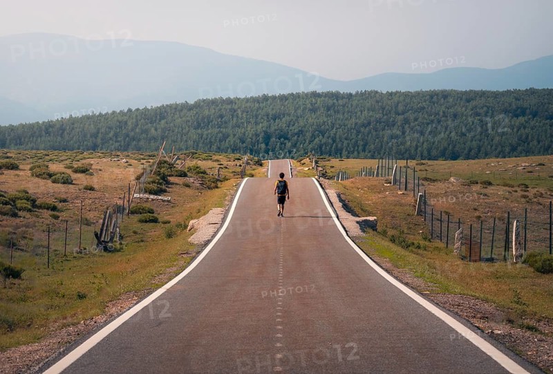 A lone man walks down a long, empty road lined with fences, surrounded by fields, heading towards the distant hills under a clear sky in Guadarrama mountain range, Madrid