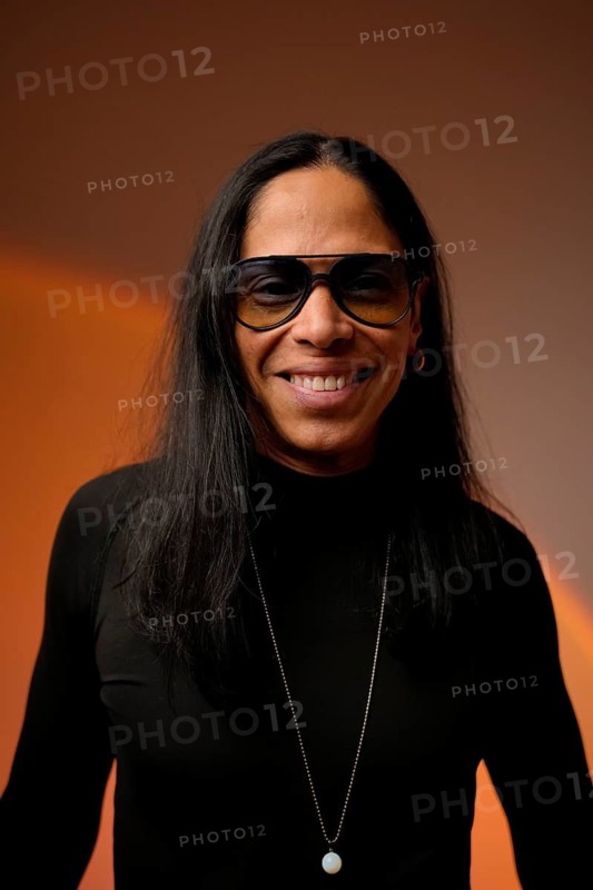 Latin man with long hair, wearing glasses and a necklace, smiles warmly. The studio background is softly lit, enhancing the relaxed and approachable vibe of the portrait