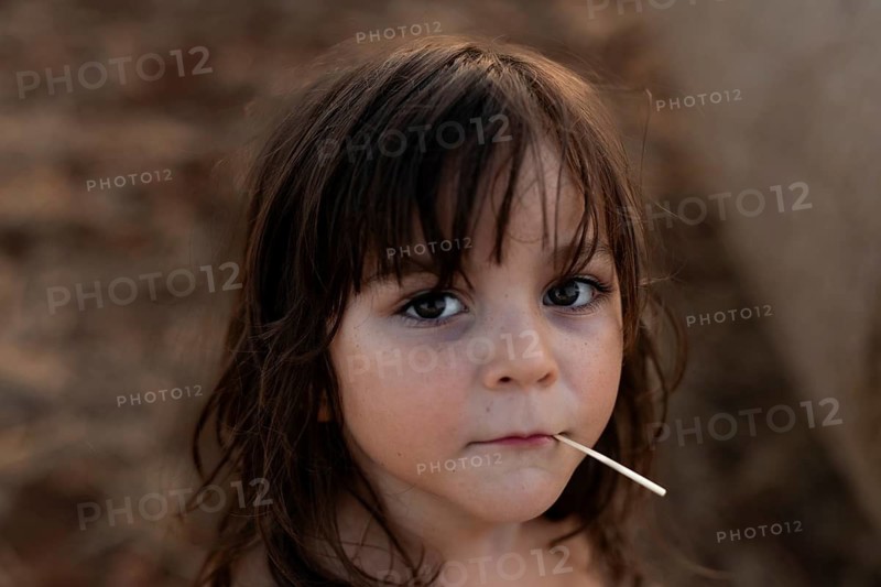 A thoughtful child with long hair gazes into the camera, holding a stick in their mouth. The soft, neutral background enhances the focus on the child's expressive eyes