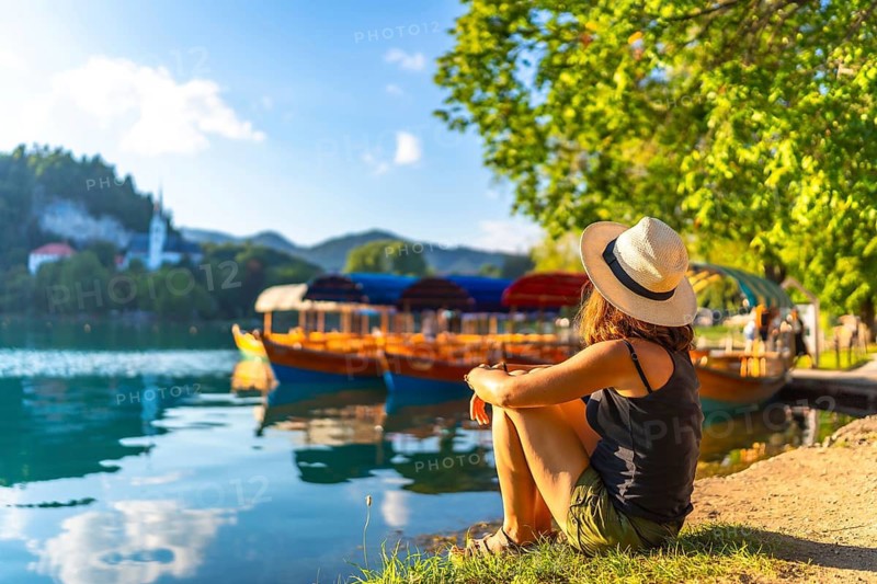 Young woman in summer attire and hat relaxing by lake bled in slovenia, enjoying views of traditional pletna boats and the church