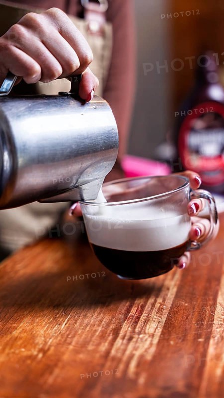 A barista artfully pours frothy milk into a glass espresso cup, creating a rich coffee blend. The scene is set on a rustic wooden table, capturing the warm cafe ambiance