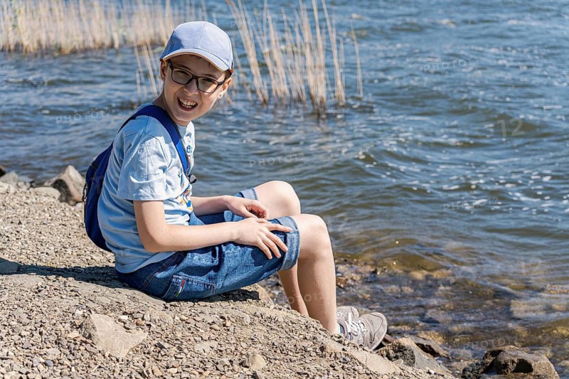 A joyful boy sits on a rocky lakeside, wearing casual summer attire and a cap, with a backpack, enjoying the sun and nature's peaceful ambiance on a bright day