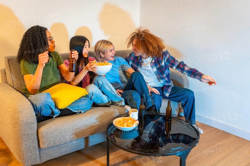 Four young friends enjoying their free time watching television, eating snacks and drinking beer, sitting on a comfortable sofa