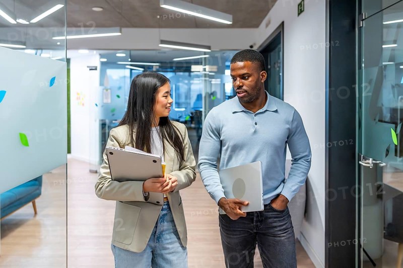 Two business colleagues are holding laptops and having a conversation while walking down an office hallway
