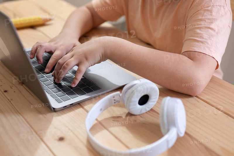 A child in a peach shirt types on a laptop at a wooden desk, with white headphones and a yellow pencil nearby, suggesting a casual learning environment