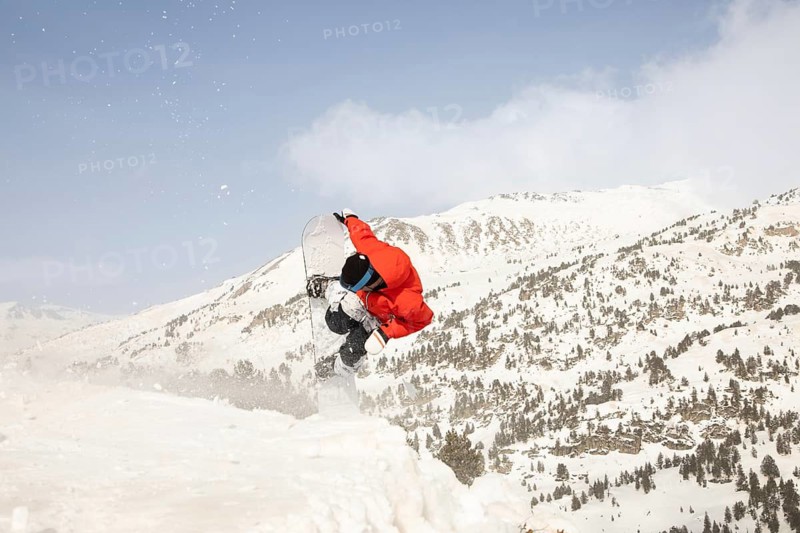 A snowboarder in a red jacket performs an impressive freeride jump amidst a stunning winter mountain backdrop, showcasing the thrill of snowboarding in fresh powder
