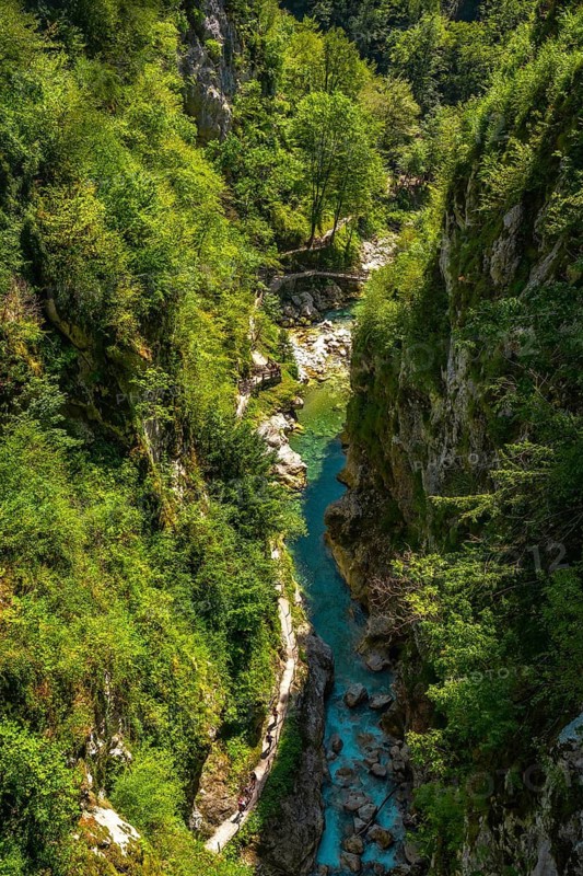 Tourists walking on a wooden footbridge over a turquoise river flowing through the tolmin gorge in slovenia, surrounded by lush green vegetation
