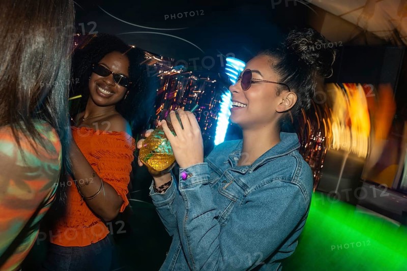 Group of diverse friends celebrating and dancing in a vibrant club, smiling and drinking cocktails while colorful neon lights create a dynamic atmosphere