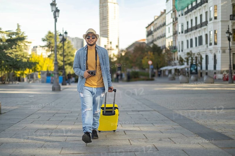 A Latino transgender man walks confidently through a city street, capturing moments with his camera, embodying pride and exploration He carries a bright yellow suitcase