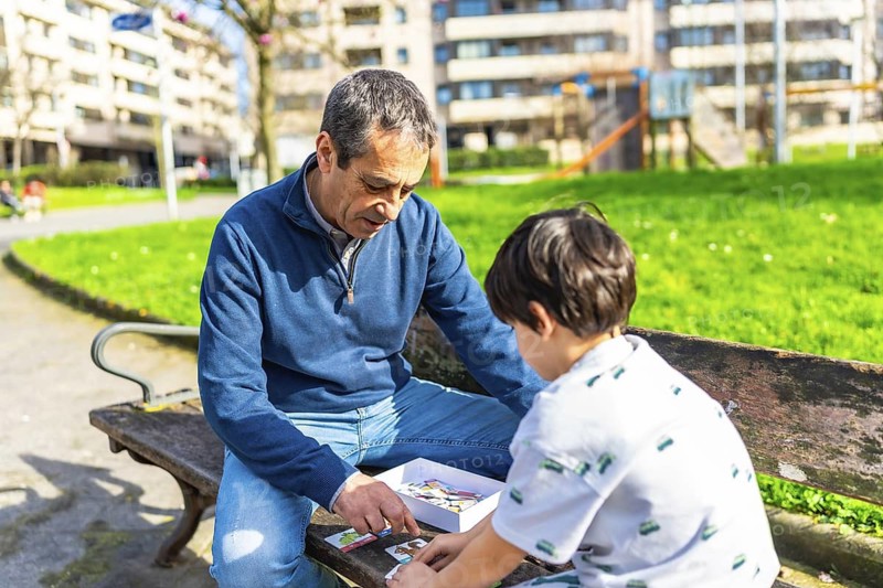 Grandfather teaching his grandson how to play cards, sitting on a bench in a park, spending quality time together on a sunny day