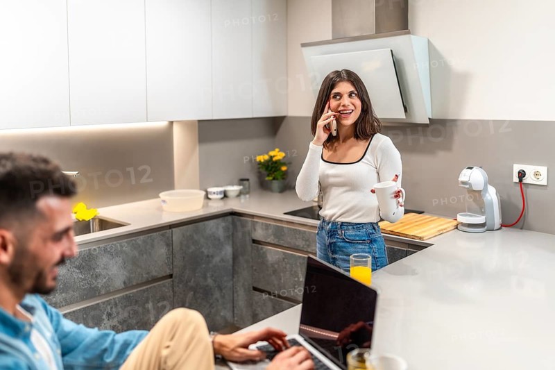 Young woman chatting on the phone while enjoying a cup, as man works on a laptop in a modern, bright kitchen, creating a cozy atmosphere