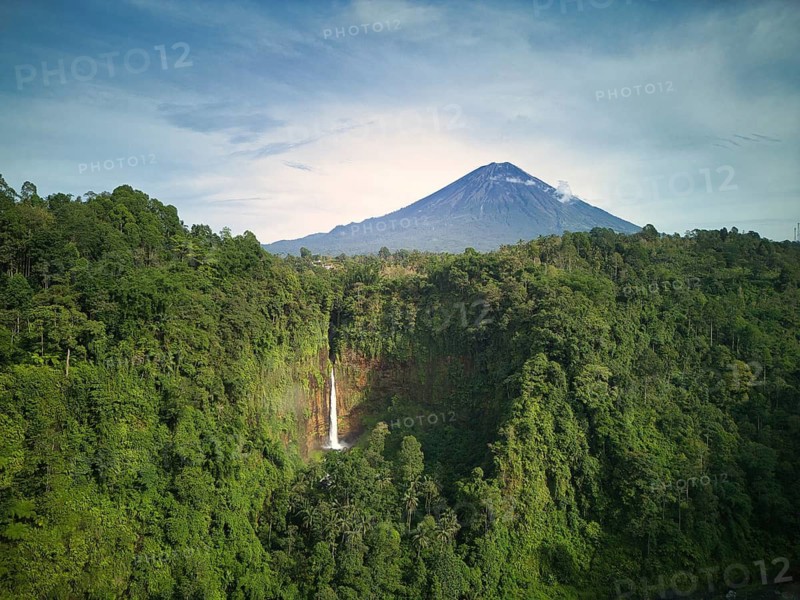 A breathtaking landscape showcasing the towering Mount Semeru volcano and the stunning Kaspa Biru Waterfall, nestled within lush green forests under a clear sky