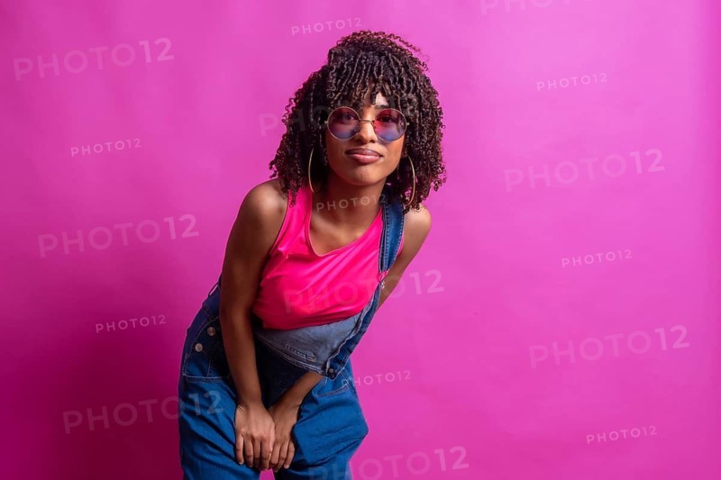 Fashionable young woman wearing sunglasses and colorful clothes posing in a studio with pink background