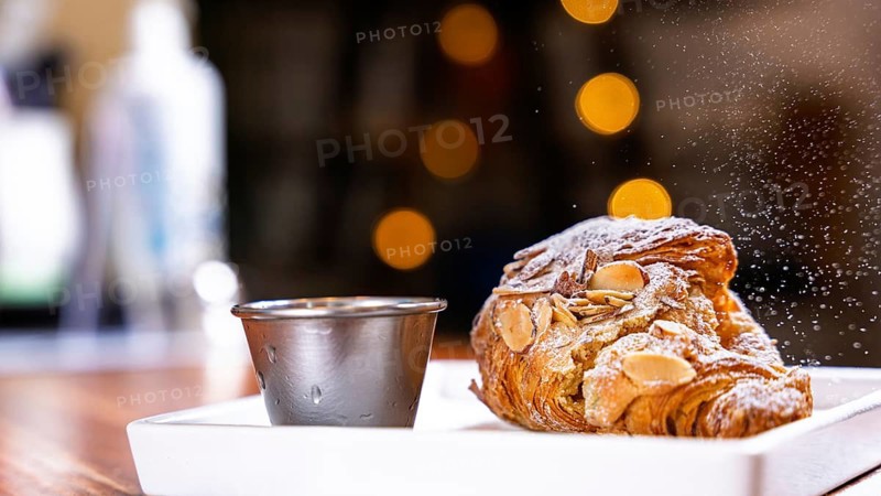 A delectable almond croissant with powdered sugar is paired with a metal cup on a cafe table. Warm bokeh lights create a cozy atmosphere, highlighting the pastry