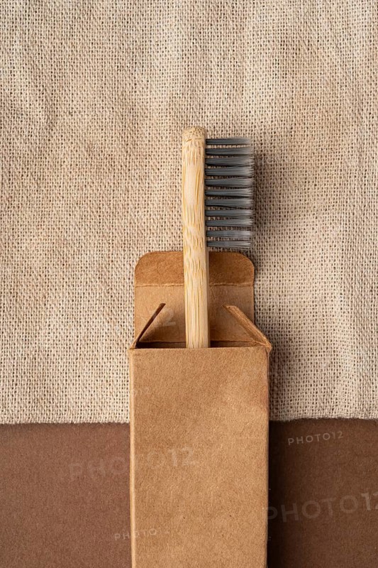 Top view of a bamboo toothbrush partially in a brown cardboard box, set against a textured beige textile background. Emphasizing eco-friendly and zero waste living