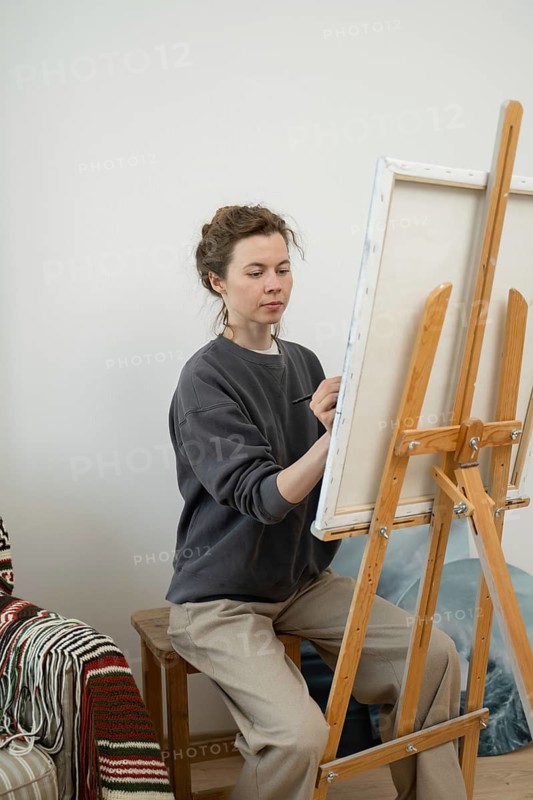 An artist focuses on creating a painting while seated at an easel in a cozy home studio. The ambiance highlights creativity and concentration in an artistic setting