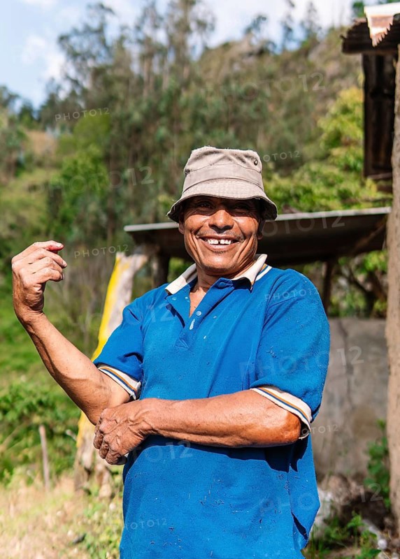 A cheerful farmer stands in a vibrant green field in Colombia, ready to collect blackberries He's wearing a blue shirt and a beige hat, enjoying the sunny day