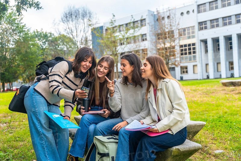 Group of female university students sitting on a concrete bench in front of a modern campus building, smiling while looking at a smartphone screen and interacting with each other