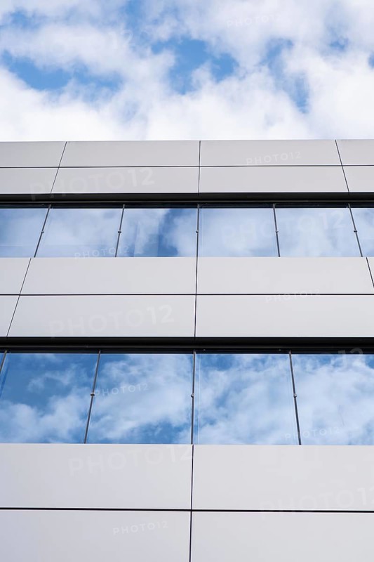A close-up view of a modern building's glass and metal facade, featuring reflective windows against a backdrop of white clouds and blue sky