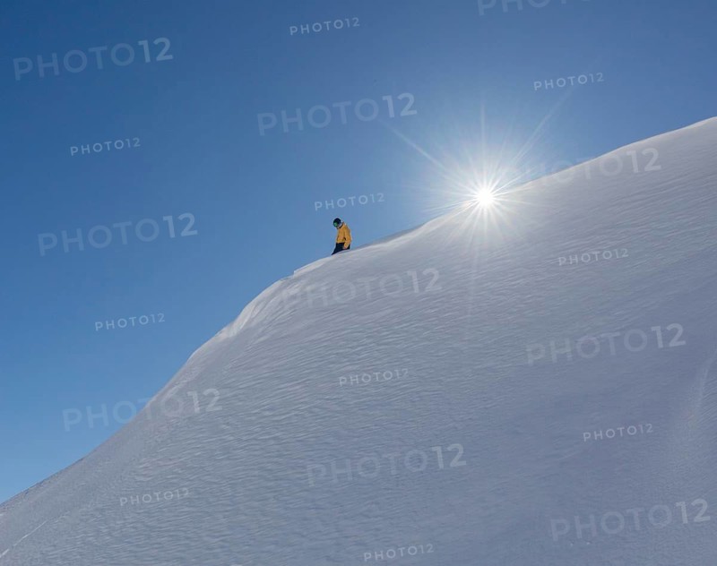 A snowboarder in a yellow jacket glides down a pristine snowy slope with the sun shining brightly above. The clear blue sky contrasts with the crisp white snow