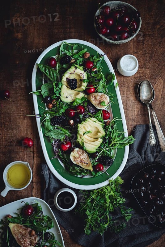 A fresh salad featuring avocado, cherries, blackberries, and figs. Served with leafy greens on a rustic wooden table, accompanied by olive oil and balsamic vinegar