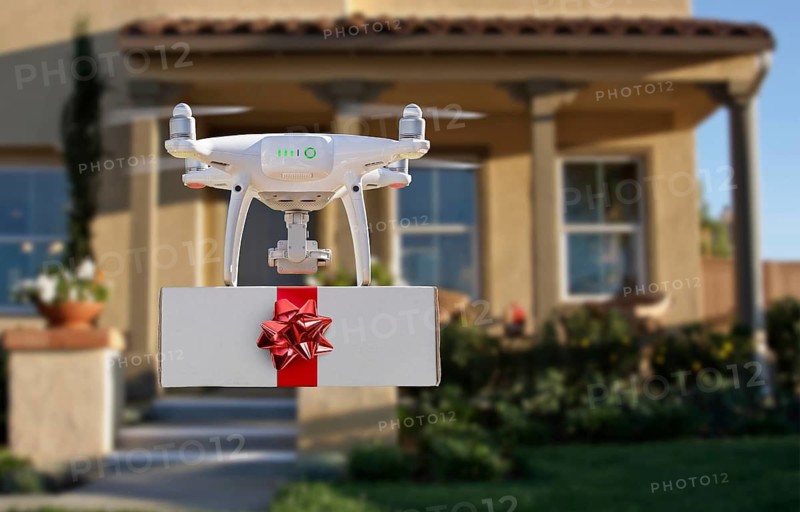 Unmanned UAV quadcopter drone delivering box with red bow and ribbon to house.