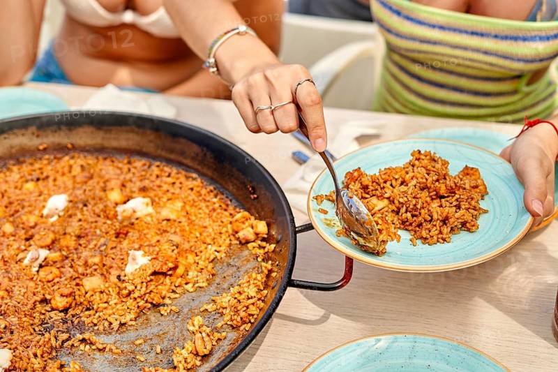 Friends gather around a large pan of paella on a sunny beach. A woman serves a generous portion onto a plate, creating a festive atmosphere perfect for summer enjoyment
