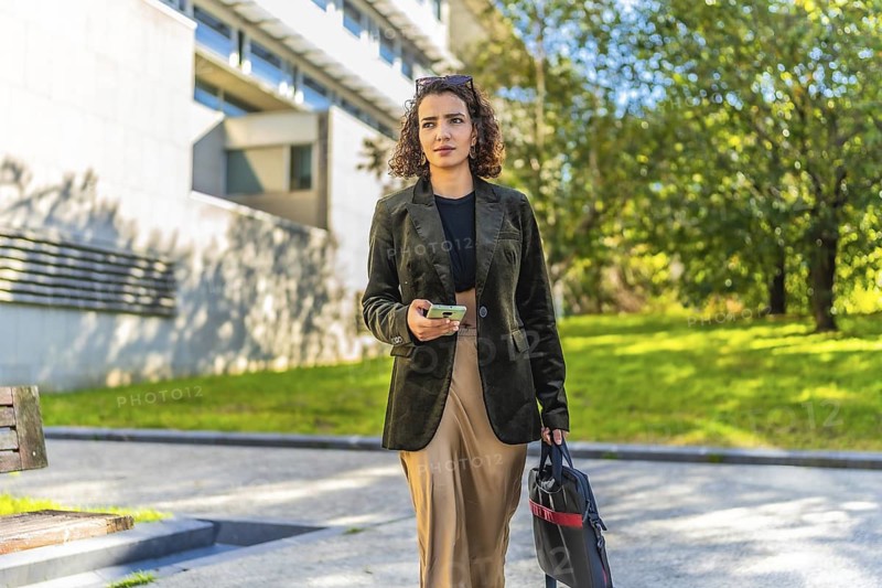Young arab entrepreneur walking along street with trees carrying laptop bag and mobile