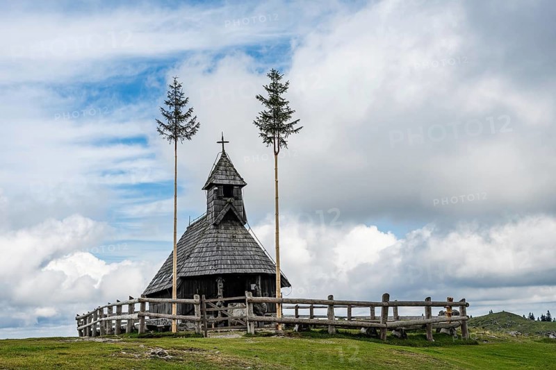 A wide mountain landscape showing a brown cow grazing in the foreground while hikers walk up a path toward a wooden chapel on a green hill in Slovenia