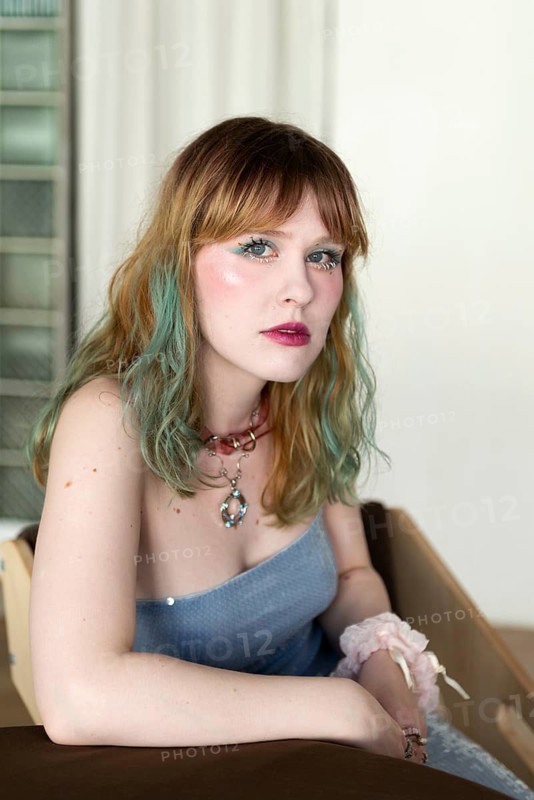 A woman with blonde and teal hair poses indoors wearing pastel pink makeup and a blue top She has a thoughtful expression, accessorized with jewelry and a scrunchie
