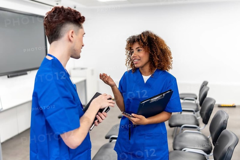 Two diverse young medical students wearing blue scrubs conversing in a modern university classroom, actively engaging in discussion and learning with clipboards in hand