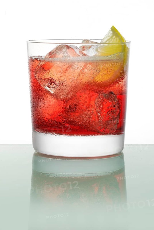 Cocktail on ice cube cubes in a tumbler on a glass table, half a slice of lemon as garnish, studio shot
