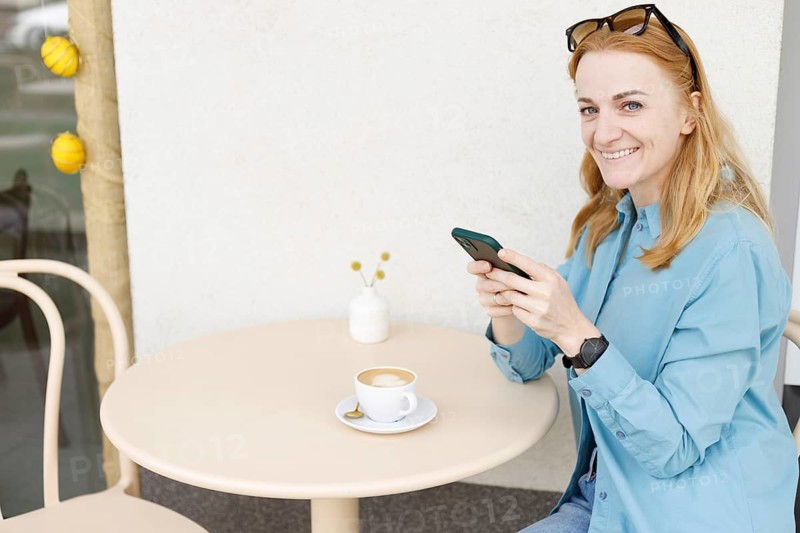 Smiling woman sitting at an outdoor cafe in Warsaw, using her smartphone and enjoying a cup of coffee Sunglasses rest on her head and a smartwatch adorns her wrist