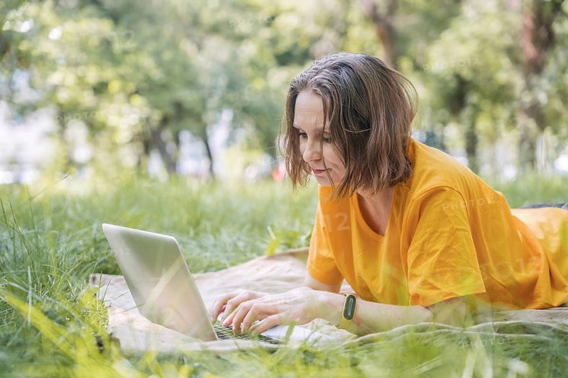 A woman lies on a blanket in a grassy park, focused on a laptop. She enjoys the outdoors while working remotely, blending nature with technology seamlessly