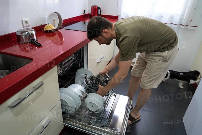A man is loading clean dishes and cutlery into a dishwasher, showcasing a typical household chore. The kitchen is modern and tidy, with a dog nearby on the floor