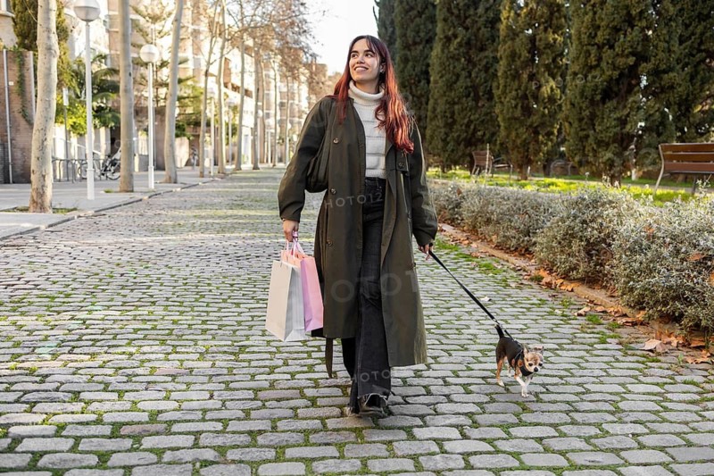 A woman walks her chihuahua on a sunny day, holding colorful shopping bags. The scene captures a relaxed moment in an urban setting with trees and cobblestone paths