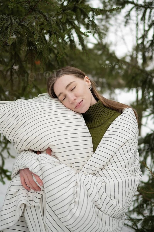 A serene scene showcasing a woman wrapped in a cozy striped blanket, embracing a pillow amidst snowy trees in the park. Her peaceful expression conveys warmth and comfort