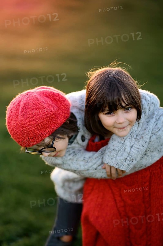 A young boy and girl, siblings, playfully embrace in a fall sunset. The boy wears glasses and a red hat, while the girl smiles warmly in a gray sweater