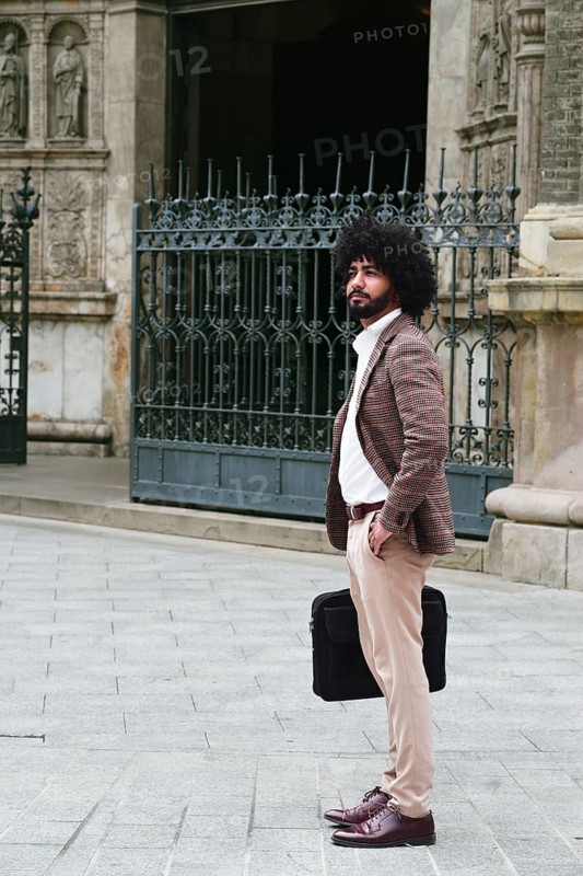 Side view of mixed-race insurance salesman with afro hair stands confidently on a stone pavement, holding a briefcase. He wears a plaid jacket, white shirt, and beige trousers. Ornamental iron gate backdrop