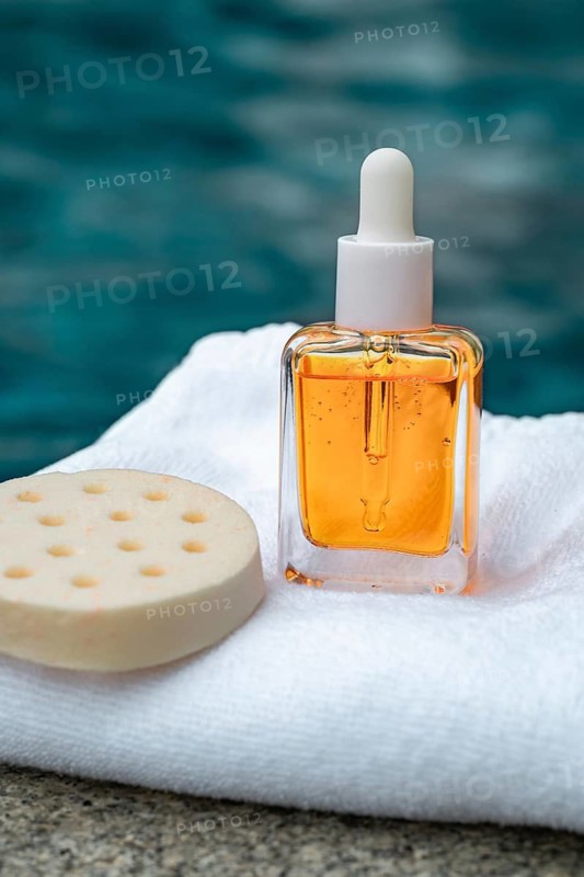 A vibrant cosmetic serum enriched with vitamins and golden flakes, captured next to a sponge on a white towel with a blurred blue sea background