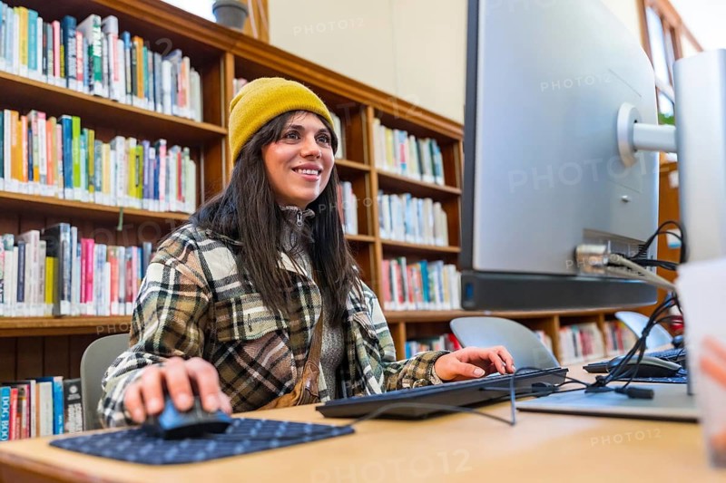 Young woman wearing a yellow beanie and plaid shirt smiling, working on a computer, and using a mouse while researching information with bookshelves in the background