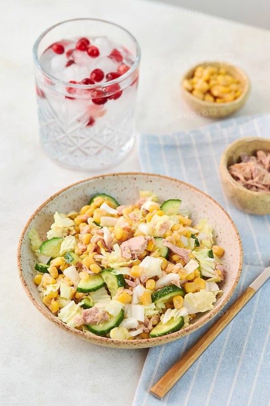 A healthy bowl of tuna salad with corn, cucumber, and cabbage on a white surface. Beside it, a glass of cold water with berries offers a refreshing touch