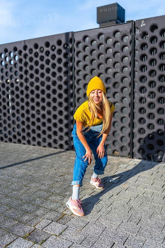 Smiling young woman wearing a yellow beanie and blue overalls dancing an urban choreography in the city